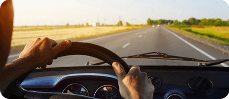 Hands gripping a steering wheel, driving on a rural road at sunset, fields on both sides.