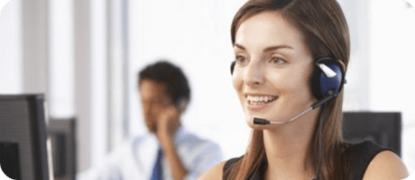 A smiling woman at a call center wearing a headset, with a colleague blurred in the background.