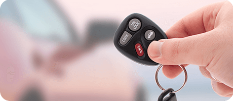 Hand holding a car key fob with lock and unlock buttons, car in the blurred background.