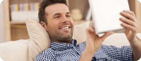Smiling man with a beard relaxing on sofa while using a tablet at home.