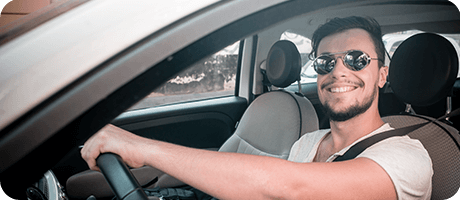 Man smiling while driving a car, wearing sunglasses and a seatbelt, in a relaxed setting.