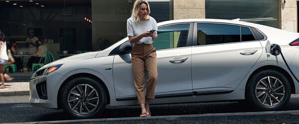 Woman standing beside a charging electric car, using a phone in an urban setting.