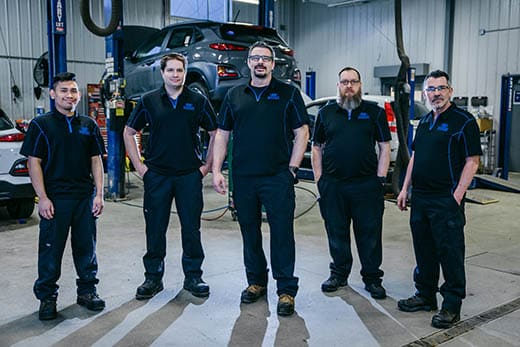 Five mechanics in a garage, wearing uniforms, with a car on a lift in the background.