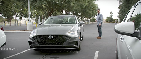 A gray Hyundai Sonata parked in a parking lot with a man standing nearby. Trees line the background.
