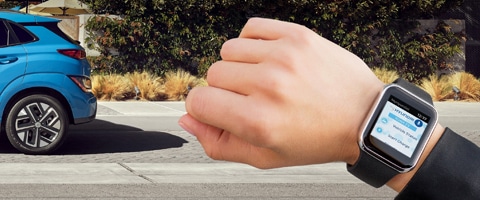 Person wearing a smartwatch controlling blue car remotely on a sunny street.