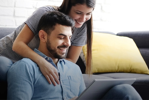 A smiling couple in casual attire looks at a tablet while sitting on a couch.