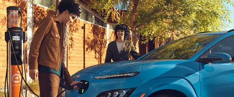 A person charges a blue electric car at a charging station, with a companion smiling nearby under a tree.