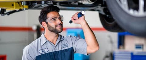 Mechanic inspecting car underside with a flashlight in a garage, wearing safety glasses.