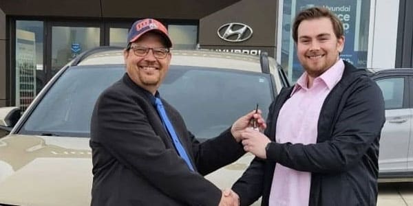 Two men shaking hands in front of a Hyundai dealership, holding car keys, symbolizing a vehicle purchase.