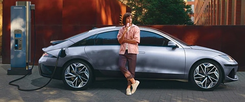 Person standing against an electric car while charging at a station, urban background.