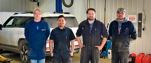 Four mechanics standing in a garage with a white SUV in the background.