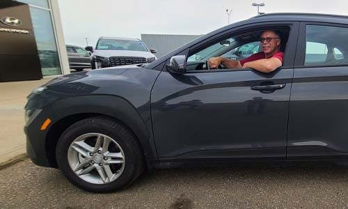 A man in a red shirt sits in a parked gray SUV outside a car dealership.