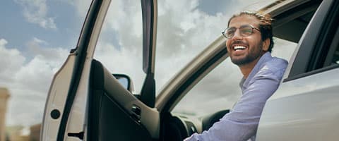 Man smiling while sitting in a car with the door open, against a cloudy sky backdrop.