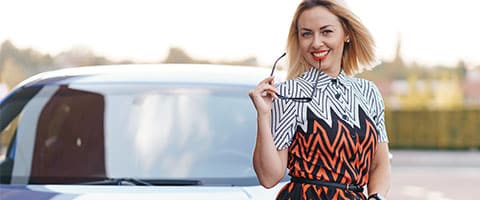 Woman smiling with sunglasses, standing beside a parked car outdoors.