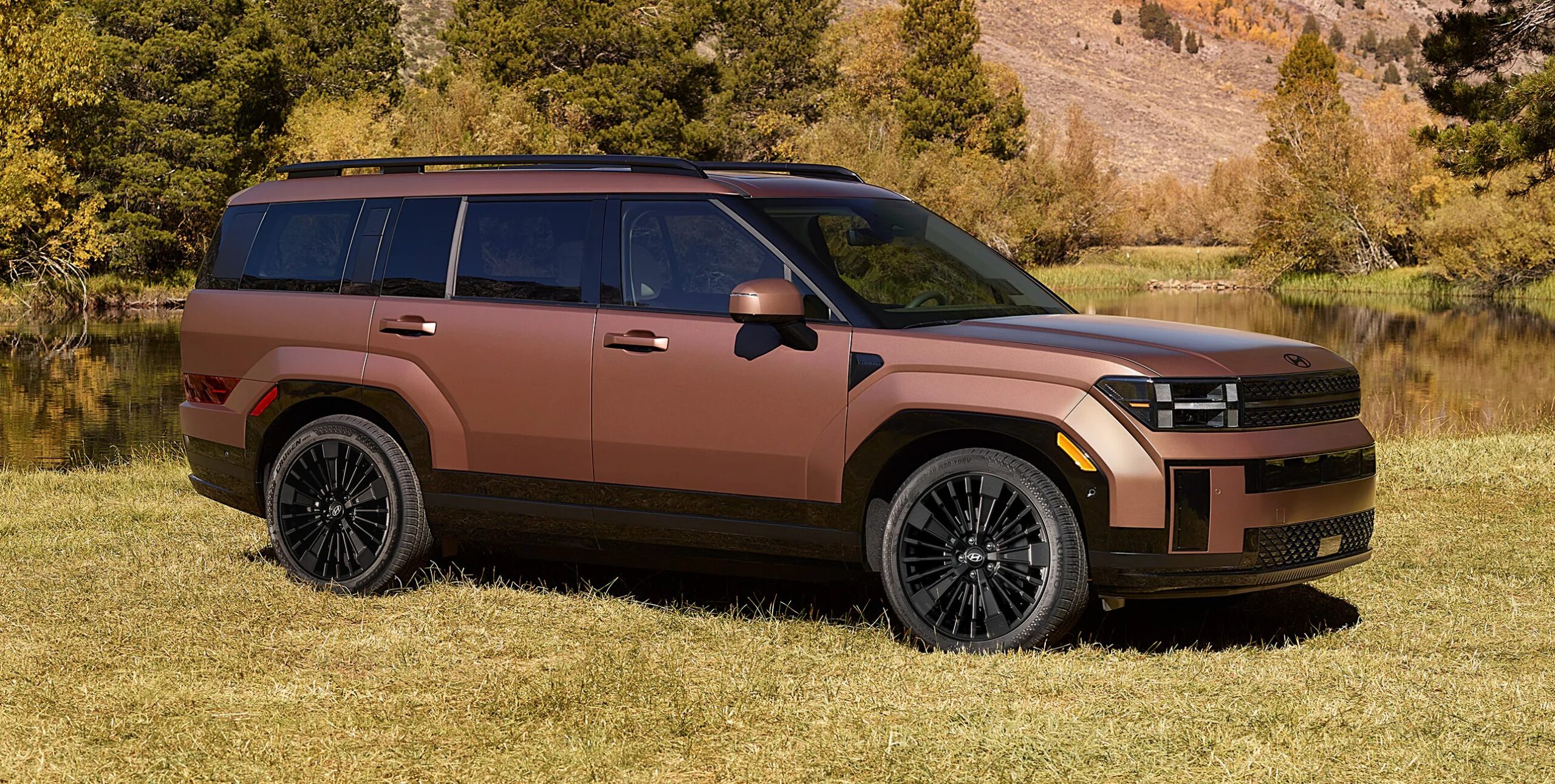 Brown SUV parked on grass near a reflective lake, surrounded by autumnal trees and mountains.